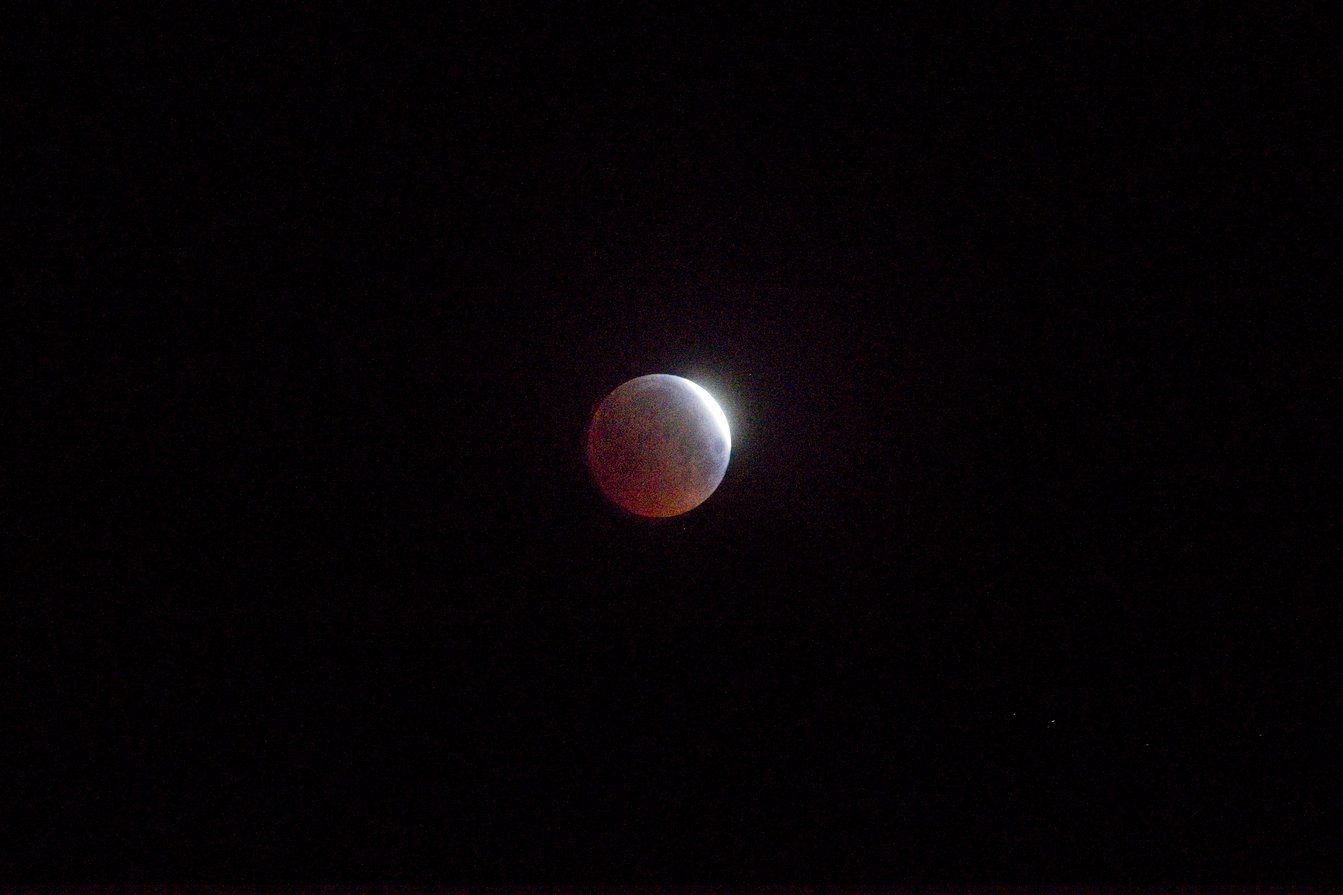 A celestial body, likely a moon undergoing a partial eclipse, set against a dark night sky. The sphere is partially illuminated with a bright, glowing white edge on one side, while the opposite portion is dimly lit with reddish-copper tones, characteristic of sunlight refracting through an atmosphere during an eclipse.